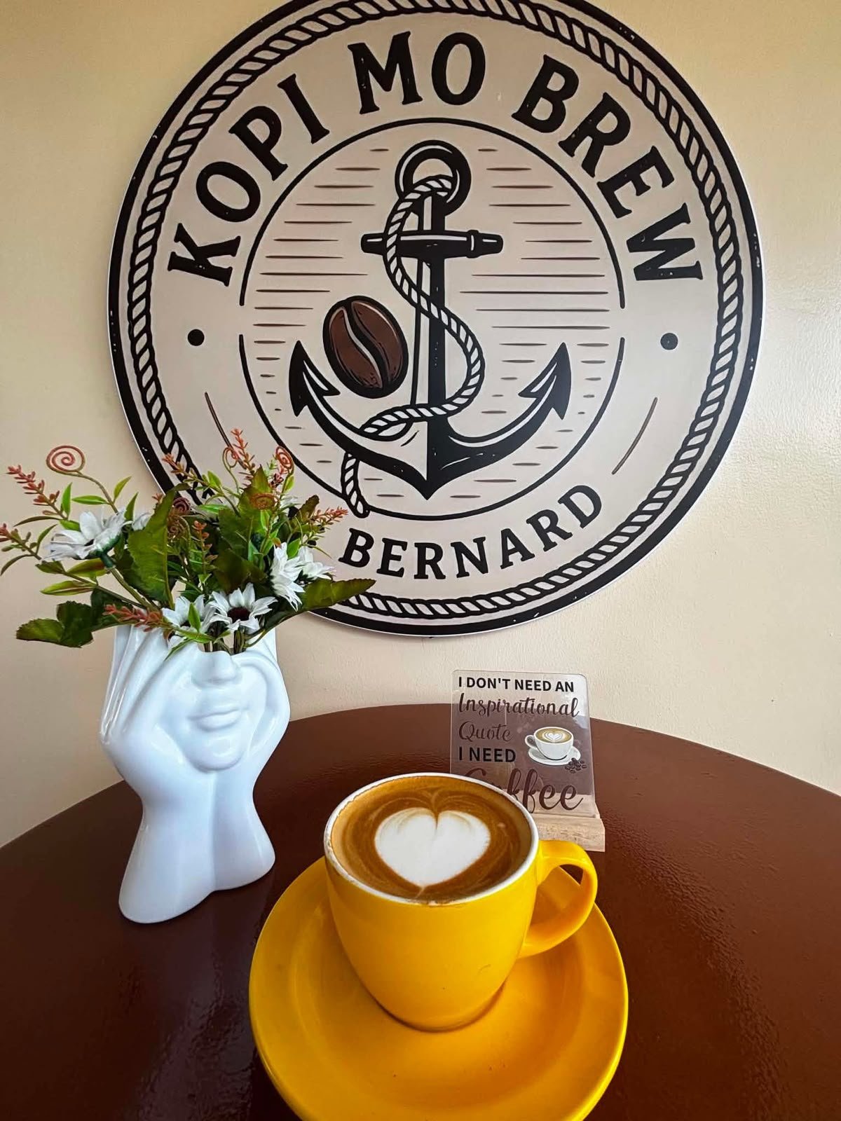 A flat white with heart latte art in a yellow cup below the Kopi Mo Brew wall sign