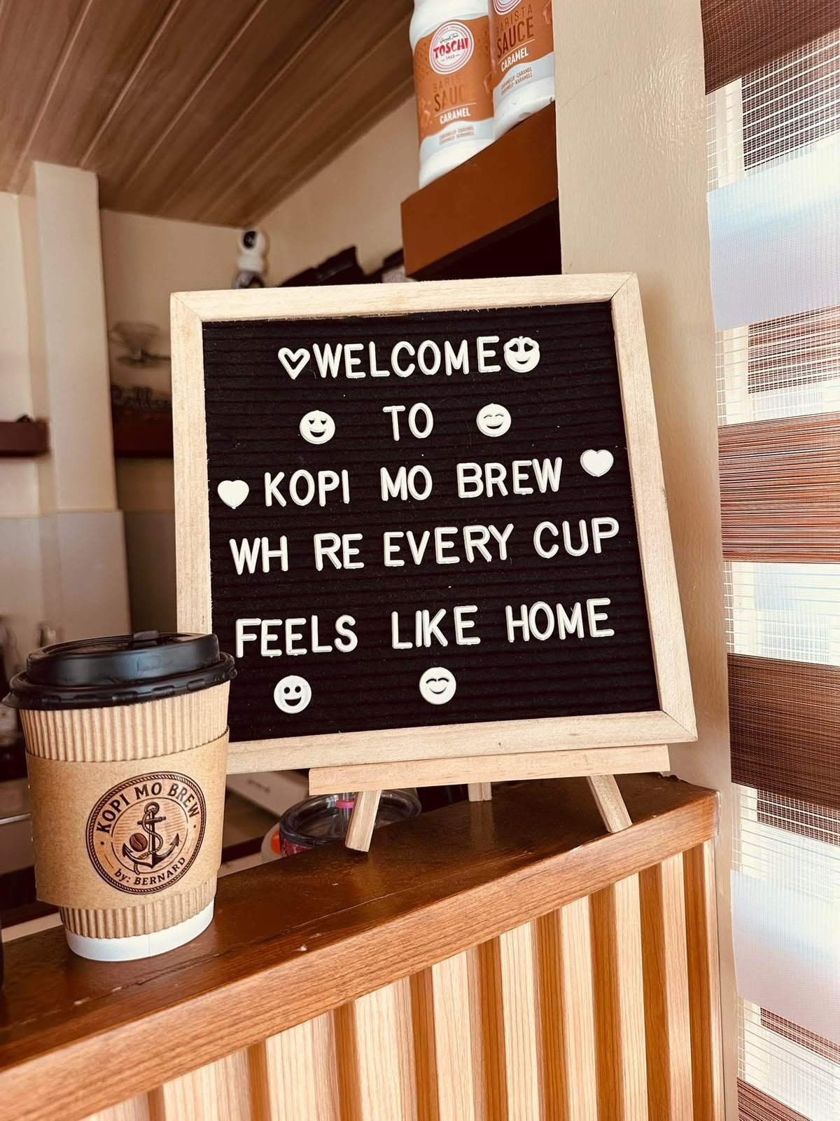 A letterboard reading Welcome to Kopi Mo Brew, where every cup feels like home, next to a kraft takeaway cup